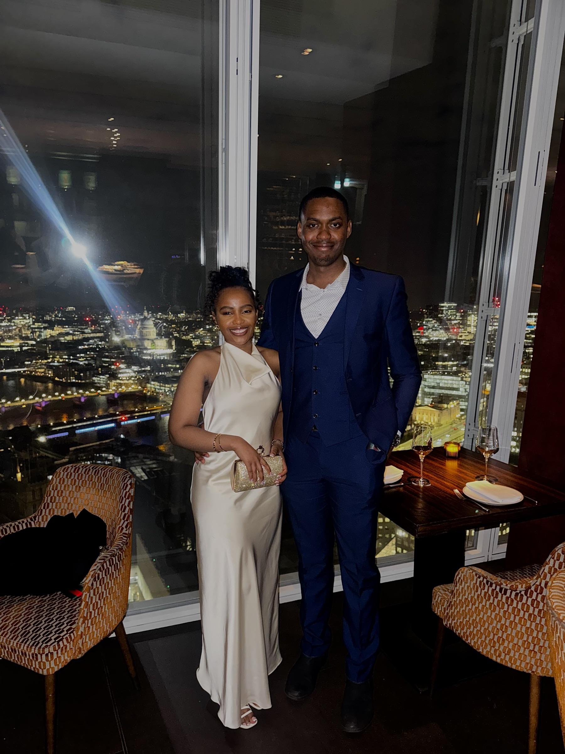 Afifoluwa and Mihlali dressed for dinner beside a restaurant window high above London, the lit city and the Thames behind them